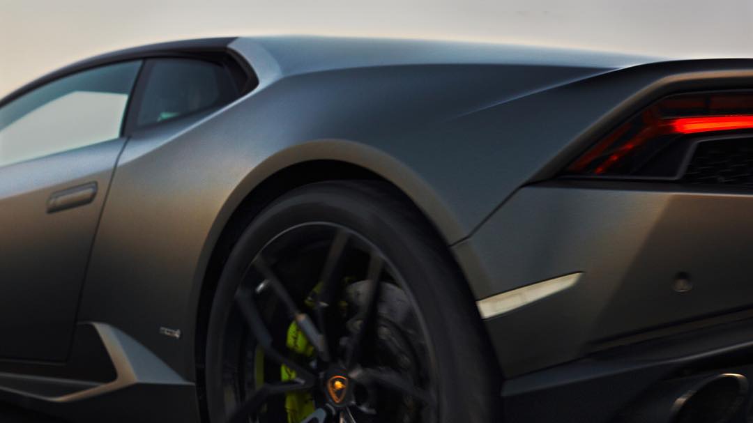 Side view of a gray Lamborghini Huracan showing the rear wheel, lights, and bodywork, set against a soft, neutral background.