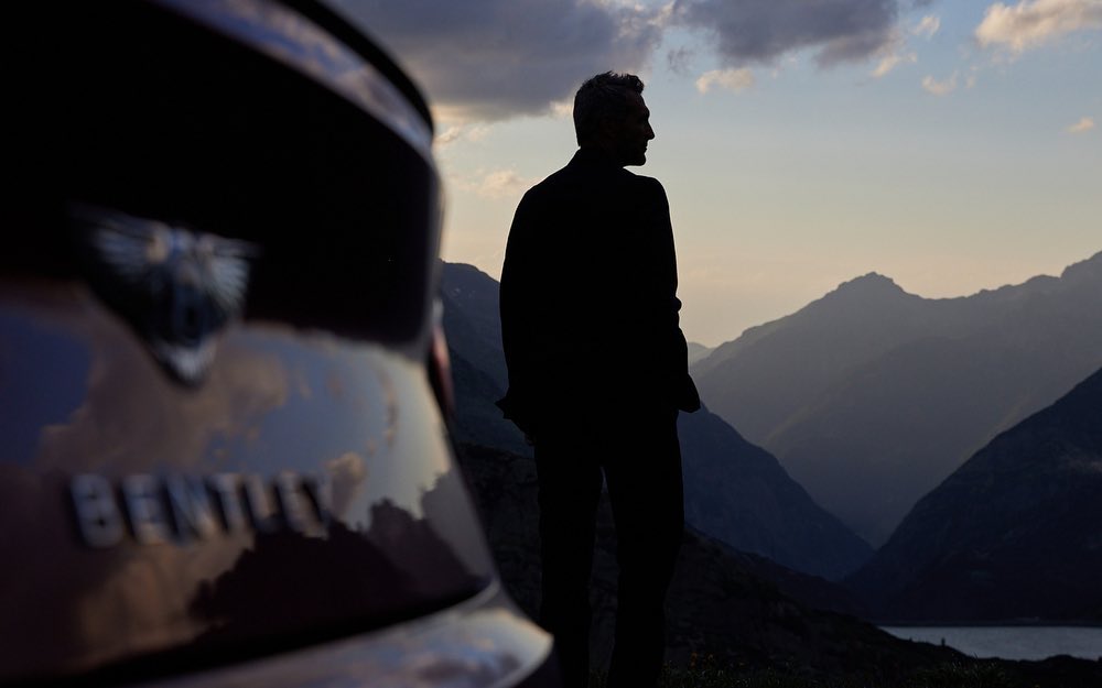Silhouette of a man standing next to a Bentley with mountains in the background at dusk, creating a scenic automotive image.