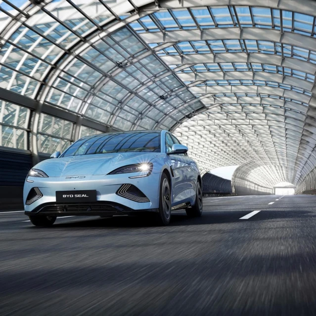 A light blue BYD Seal electric car drives through a modern, transparent tunnel with sunlight visible in the distance.