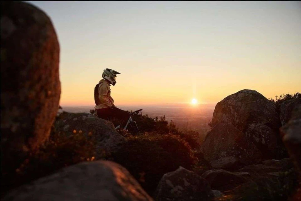 A man in a helmet sits on a mountain bike on a rocky hilltop, silhouetted against the rising sun in a scenic outdoor landscape.