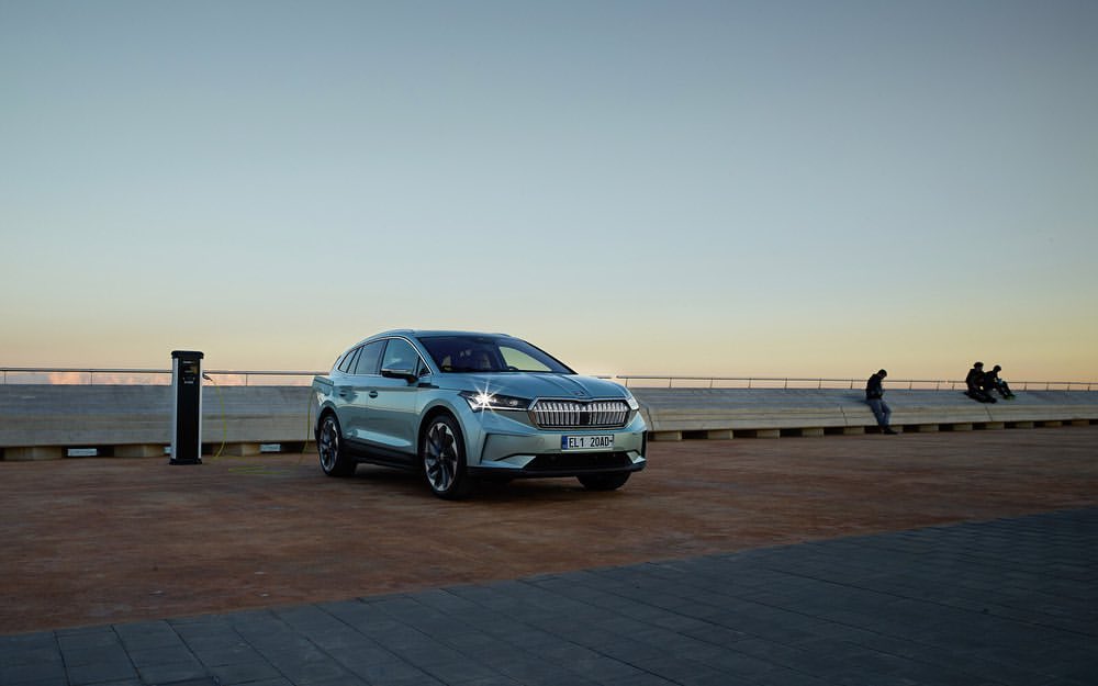 Silver electric SUV parked at a charging station near a waterfront promenade with people silhouetted in the background at dusk.