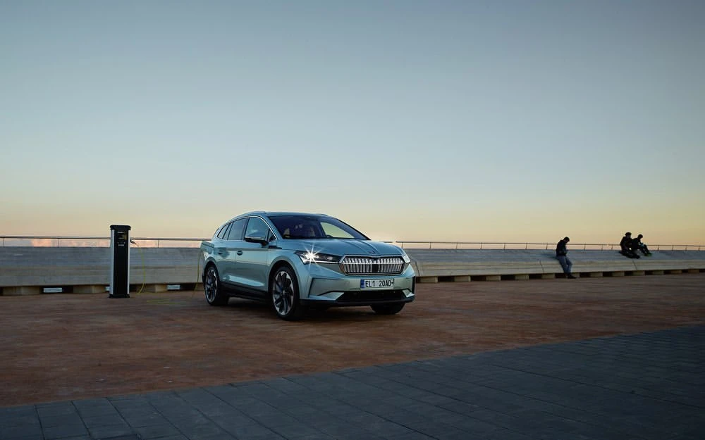 Silver electric SUV parked at a charging station near a waterfront promenade with people silhouetted in the background at dusk.