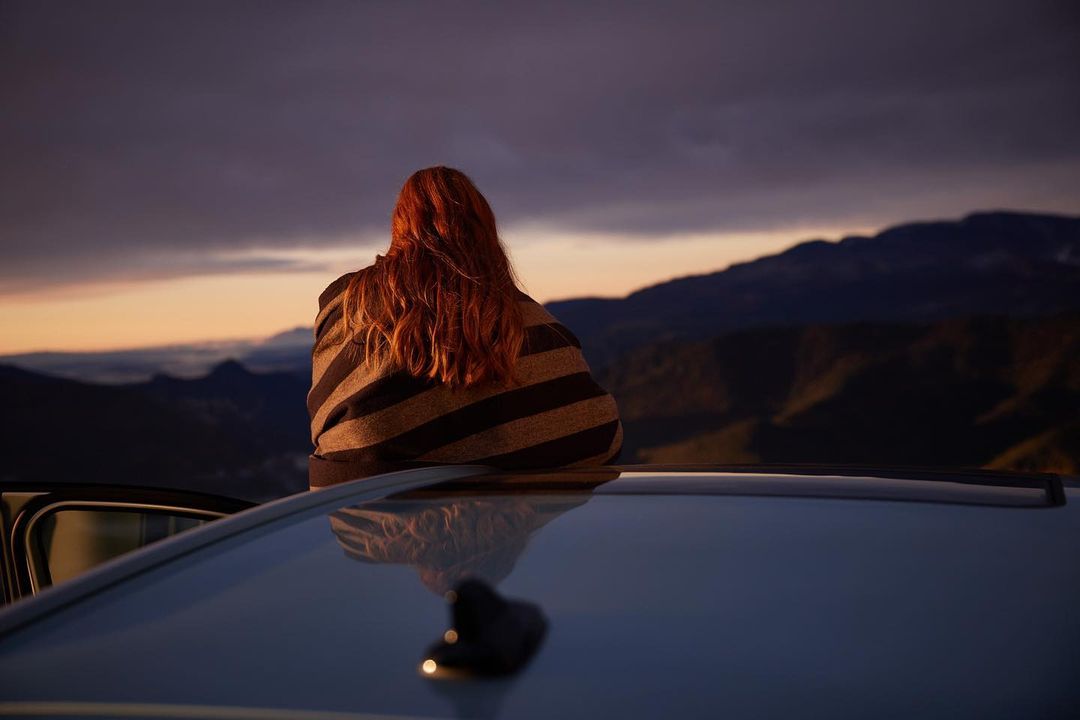 Rear view of a red-haired woman wrapped in a striped blanket sitting on a car roof at sunset
