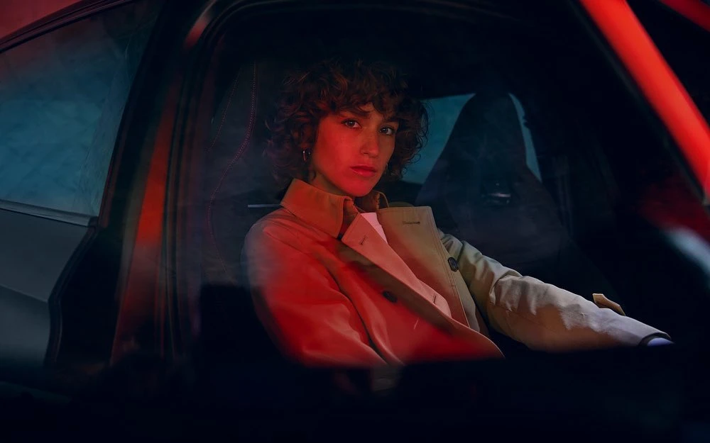 Woman with curly hair sits inside a car, illuminated by red lighting at night, wearing a tan trench coat and a white shirt.