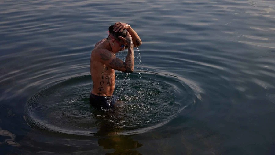Tattooed man in a lake, wearing swimming goggles, with hands on his head and water dripping, captured in an eye-level outdoor shot.