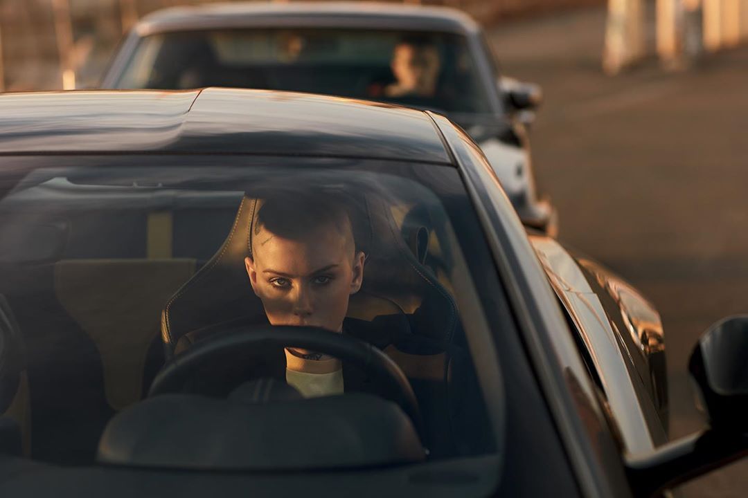 Woman with a shaved head driving a luxury car, with another car visible in the background. Automotive-themed imagery.