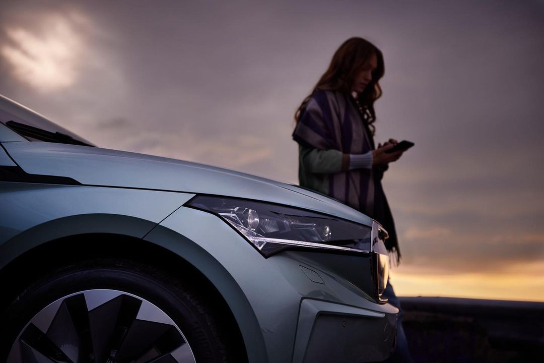 A light green Skoda car parked on a roadside at sunset with a female adult using a smartphone visible next to the vehicle.