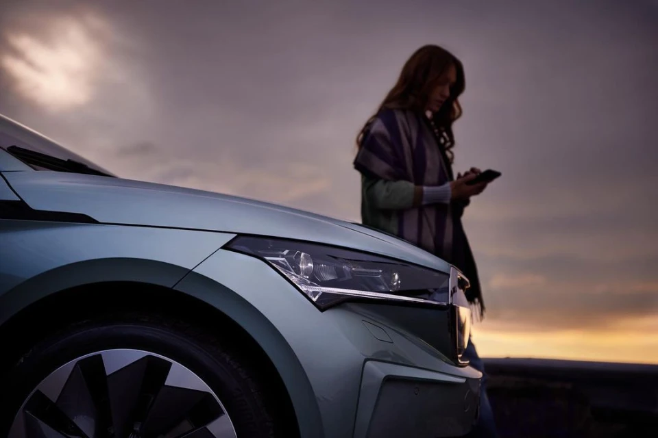 A light green Skoda car parked on a roadside at sunset with a female adult using a smartphone visible next to the vehicle.