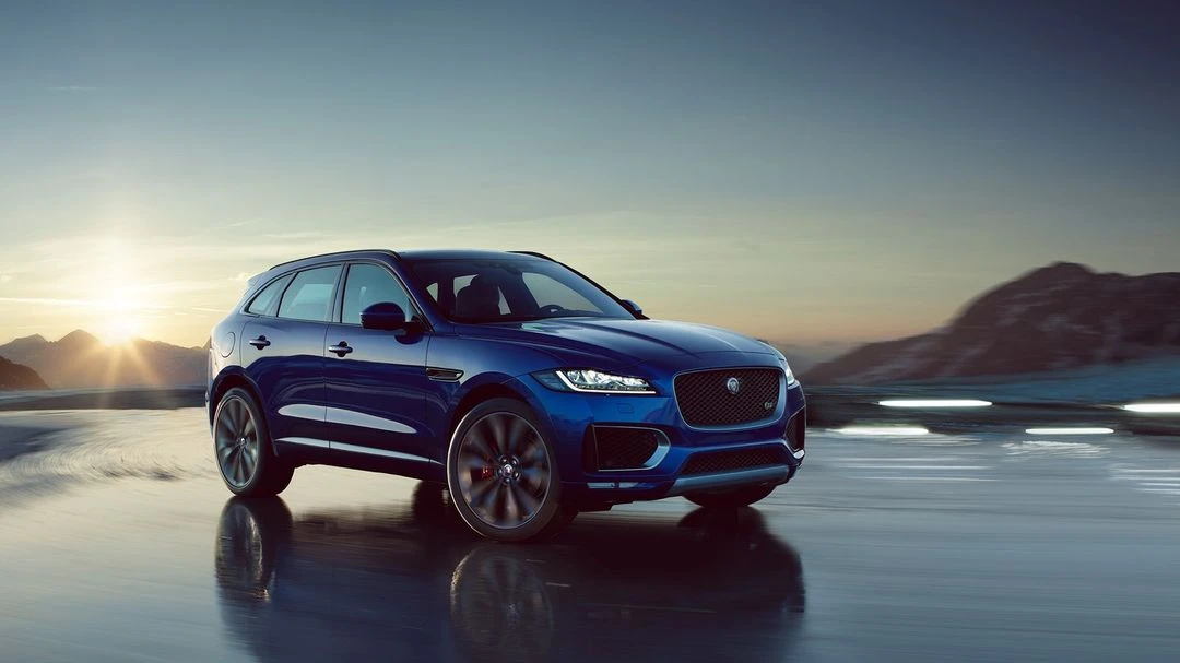 Blue Jaguar F-Pace SUV parked on a wet surface with mountain range and sunset in the background, capturing a luxury automotive moment.
