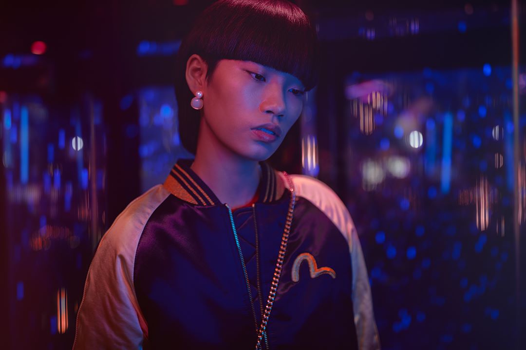Stylish Asian woman in blue satin bomber jacket, gold chain, and pearl earrings posing in an atmospheric nightclub setting with neon lights.