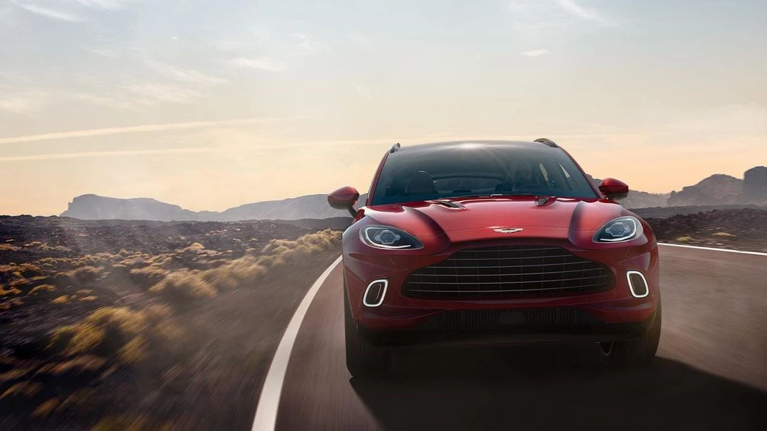 Red Aston Martin SUV driving on a highway with a desert mountain range under an open sky. Automotive beauty shot with motion blur.