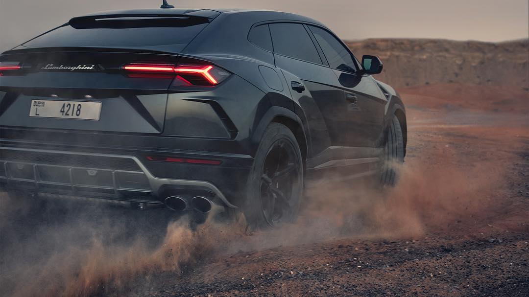 A dark grey Lamborghini Urus SUV kicks up dust on a reddish-brown desert landscape in a promotional shot, partially angled from the rear.