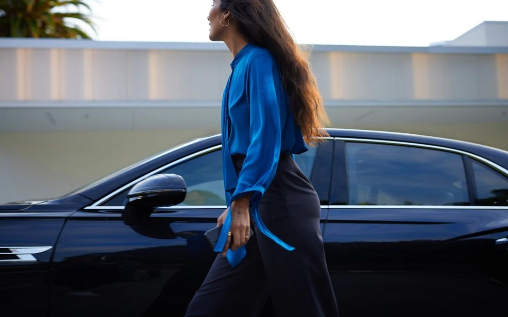 Stylish woman in blue blouse walking by a black sedan. She's holding a mobile phone with a modern building and palm trees in the background.