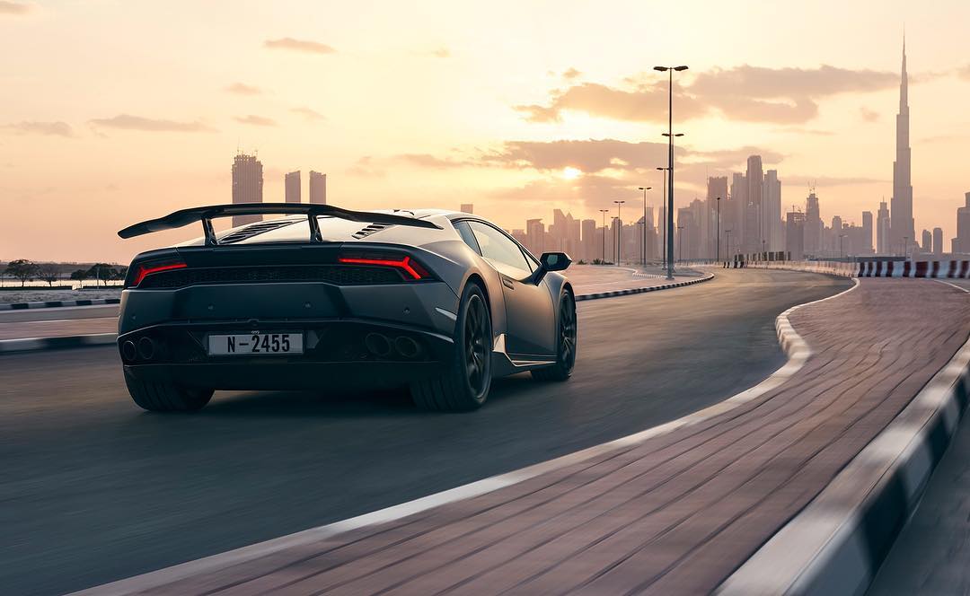 Sleek black Lamborghini racing down a paved road with the modern Dubai skyline in the background at sunset. Automotive photography.