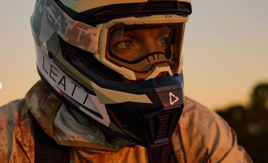 Close-up shot of a person wearing a LEATT camouflage motocross helmet and goggles, focusing on the helmet's design and the rider's eyes.