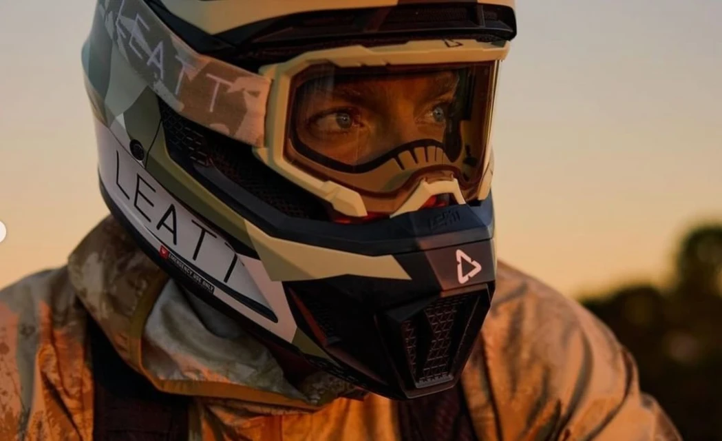 Close-up shot of a person wearing a LEATT camouflage motocross helmet and goggles, focusing on the helmet's design and the rider's eyes.