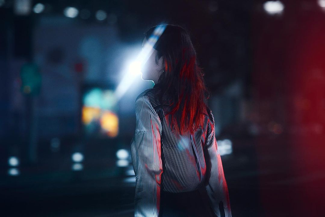 Woman in striped satin shirt standing outdoors at night with dramatic lens flare across her face