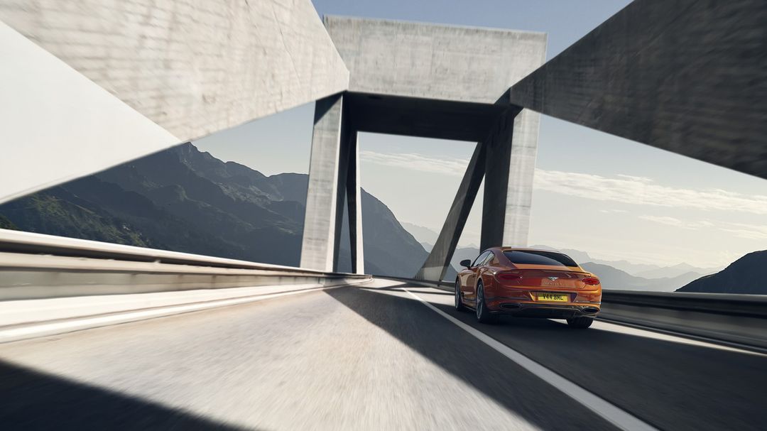 Orange car on a highway under a concrete bridge against a mountain backdrop on a clear day, capturing speed and luxury.