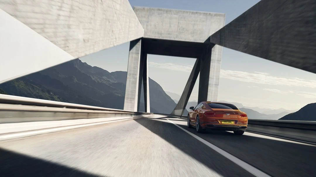 Orange car on a highway under a concrete bridge against a mountain backdrop on a clear day, capturing speed and luxury.