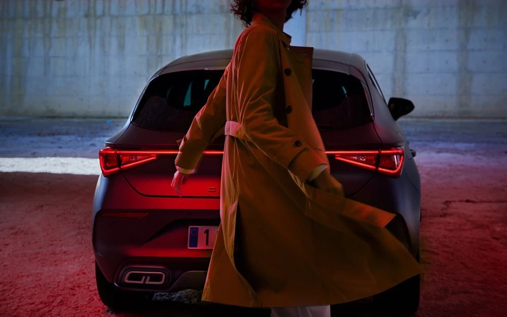 Woman in trench coat stands near a dark grey car with glowing red taillights in an indoor setting, lit with red and blue.