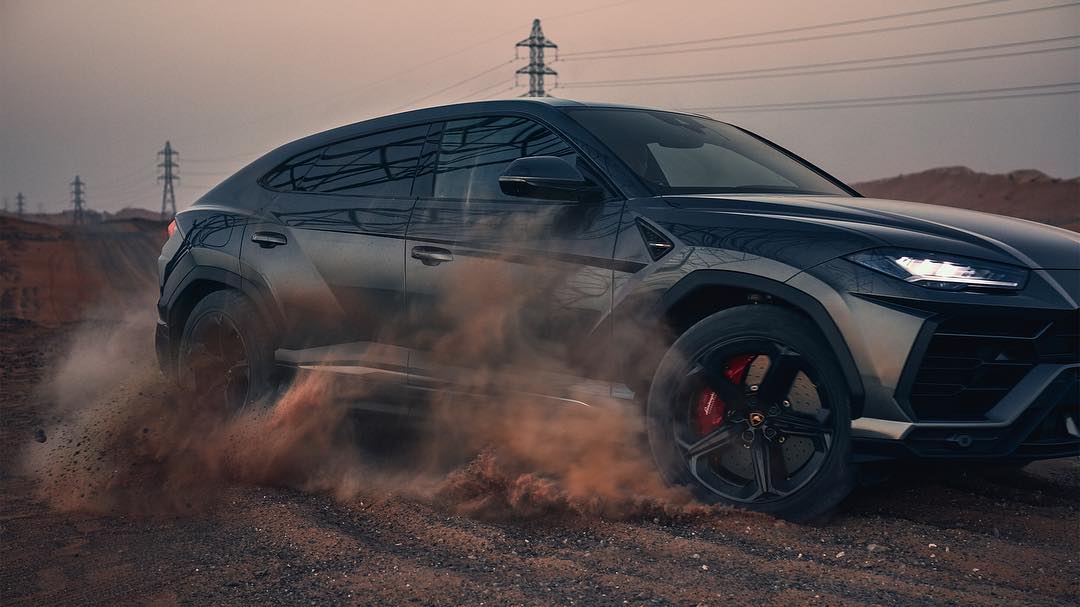 Luxury black Lamborghini Urus SUV speeds across a dirt road, kicking up dust clouds with transmission towers in the background.