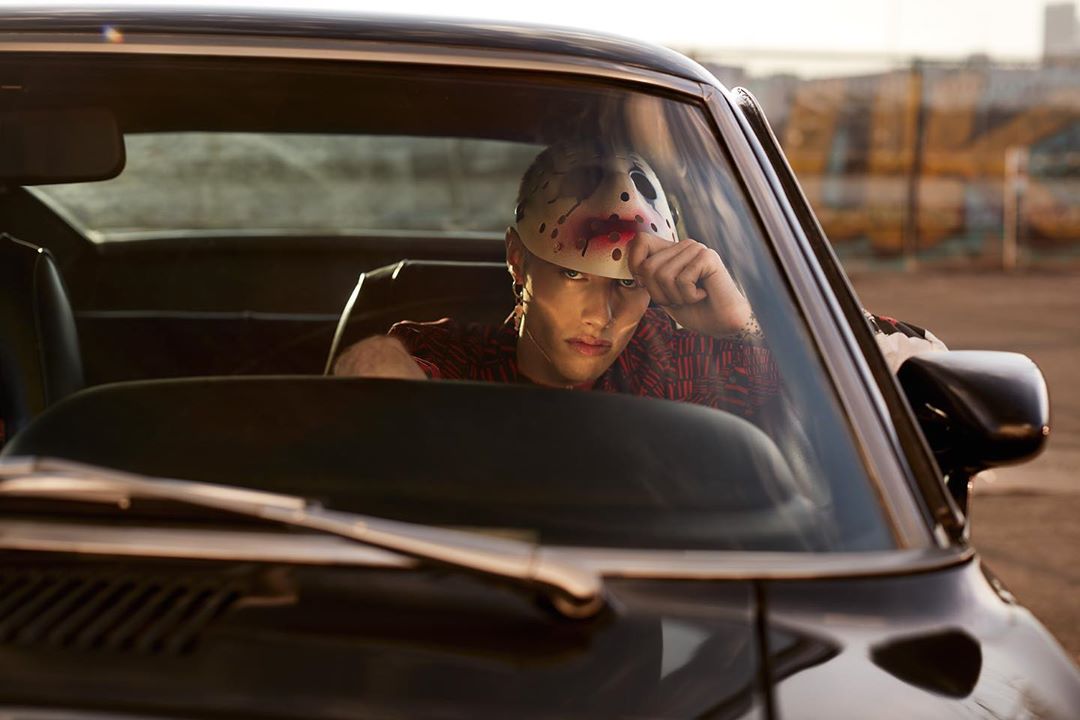 Person in hockey mask driving a black car. Dark vehicle in an urban outdoor setting, with sunlight illuminating the scene.