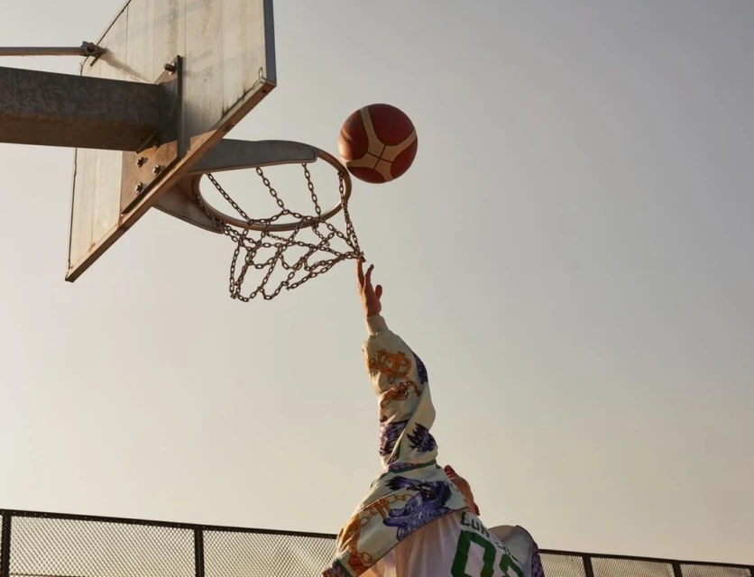 Low-angle view of a person reaching for a basketball by a chain net and metal hoop against a clear sky, capturing an athletic moment.