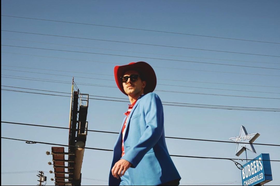 Adult male in cowboy hat and blue suit poses outdoors near roadside Astro Burgers sign against a clear blue sky on a sunny day.