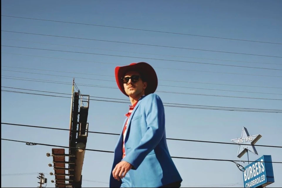 Adult male in cowboy hat and blue suit poses outdoors near roadside Astro Burgers sign against a clear blue sky on a sunny day.