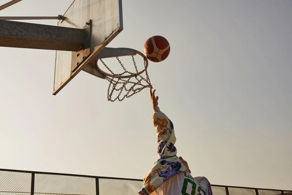 Action shot of a basketball player reaching for the ball near a hoop with a chain net under a bright sky in an outdoor setting.