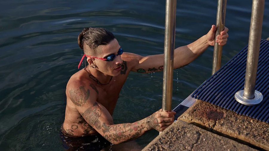 Tattooed man with swimming goggles emerges from ocean holding onto metal ladder; ripples in water surface.