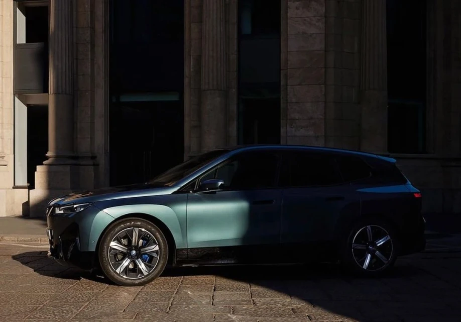 Side view of a teal BMW iX electric SUV parked on a cobblestone street in front of a light stone building with arched windows.