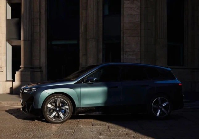 Side view of a teal BMW iX electric SUV parked on a cobblestone street in front of a light stone building with arched windows.