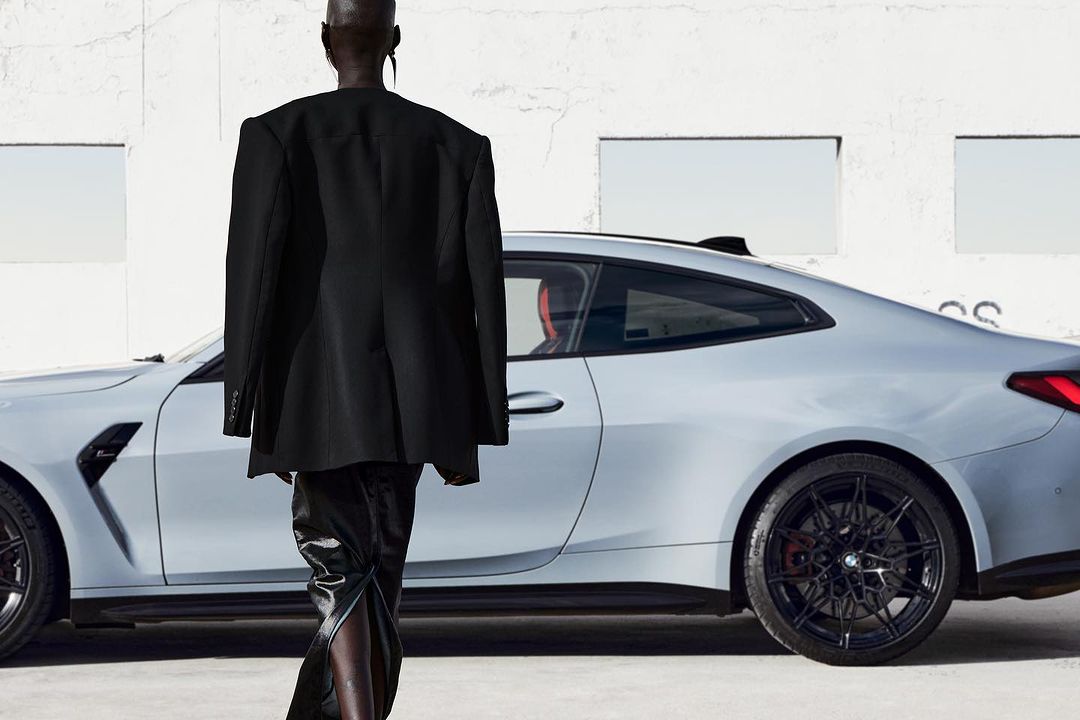 Fashion shoot showcasing a person in a black blazer and pants walking in front of a light gray BMW coupe against a white building.