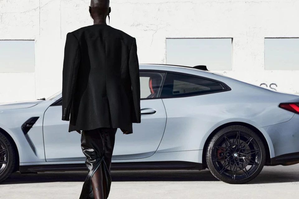 Fashion shoot showcasing a person in a black blazer and pants walking in front of a light gray BMW coupe against a white building.