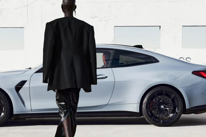 Fashion shoot showcasing a person in a black blazer and pants walking in front of a light gray BMW coupe against a white building.
