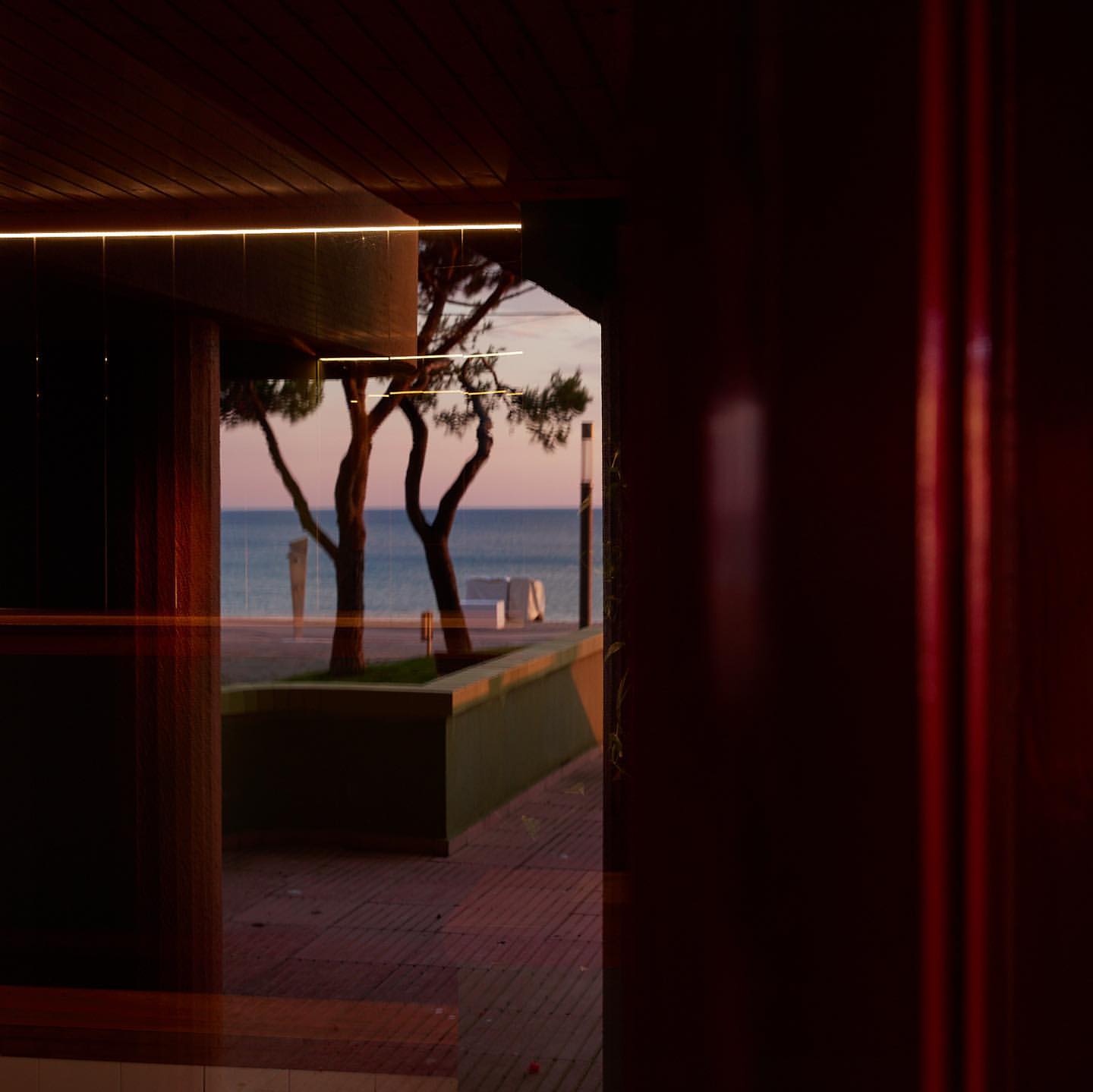 Dusk view through window of tranquil ocean scene with trees, walkway, and seating, showcasing serene colors of twilight sky and sea.