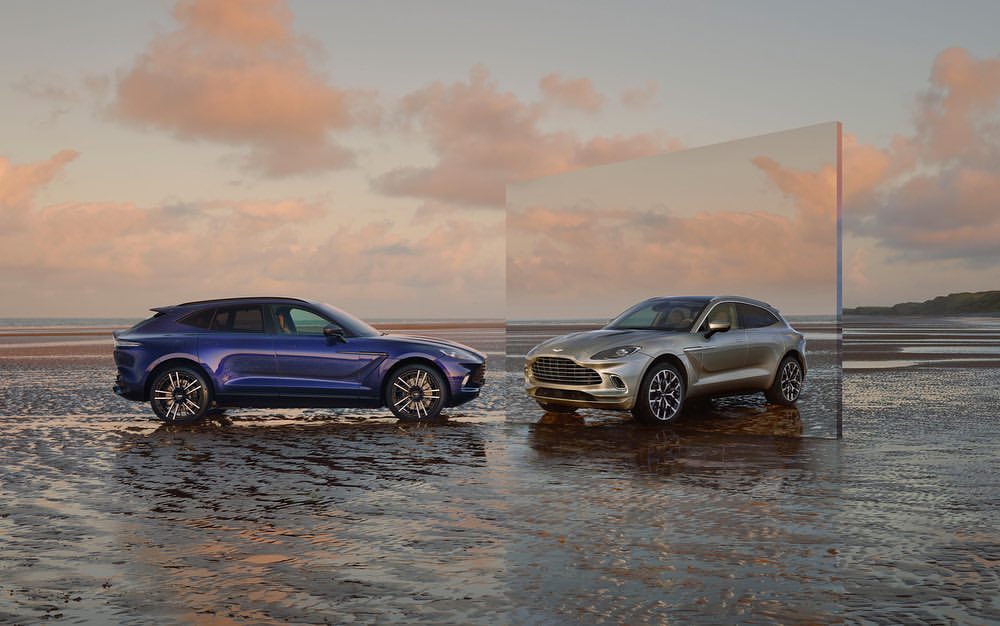 Two Aston Martin DBX SUVs, one blue and one silver, parked on a wet beach at sunset with a large mirror reflecting the beach.