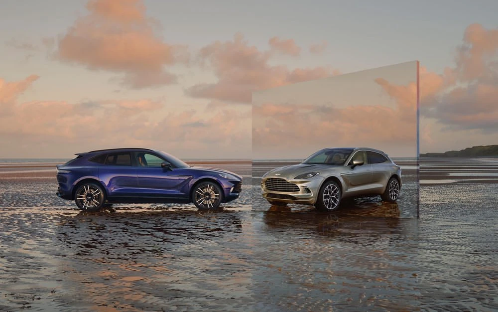 Two Aston Martin DBX SUVs, one blue and one silver, parked on a wet beach at sunset with a large mirror reflecting the beach.