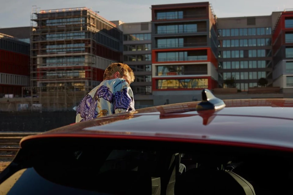 Person stands near a red car in front of modern apartment buildings. Man in patterned shirt looks at urban scenery in the evening light.