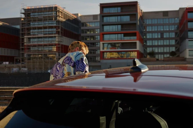 Person stands near a red car in front of modern apartment buildings. Man in patterned shirt looks at urban scenery in the evening light.
