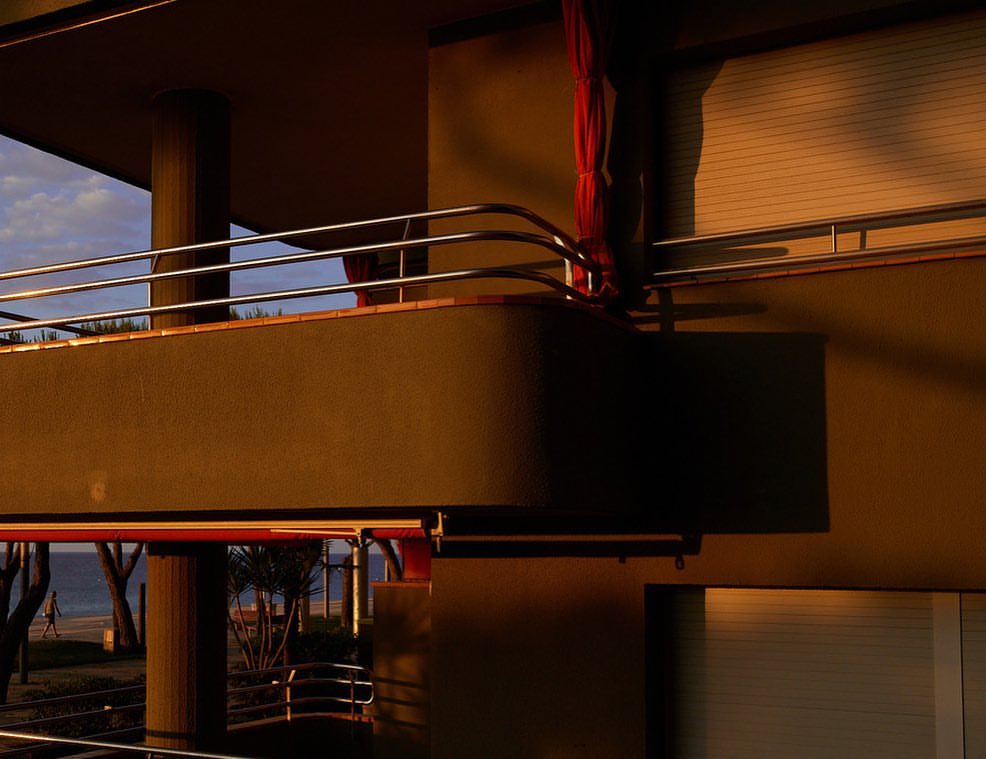 Exterior view of modern architecture features an elevated balcony with metal railing, columns, and warm golden sunlight.