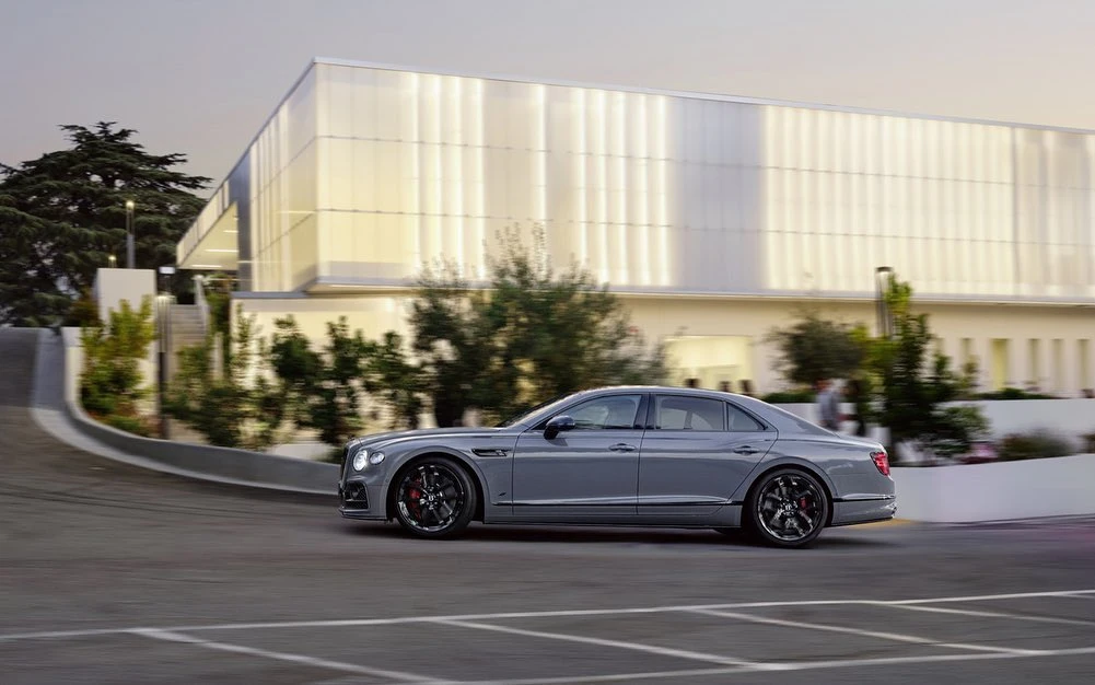 Sleek gray Bentley sedan driving on asphalt with a modern building in the background, emphasizing luxury automotive design and urban scenery.