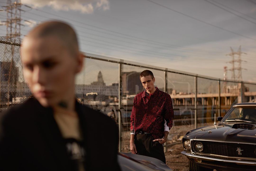 Two models pose outdoors by a black car and a chain link fence, with a cityscape and power lines visible under a cloudy sky.
