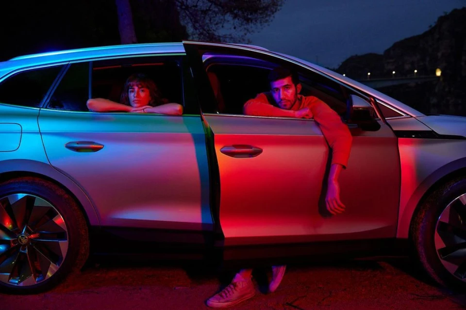 Silver crossover SUV with two people at night. Man leans from the driver's seat; woman sits in the back. Automotive product shot.