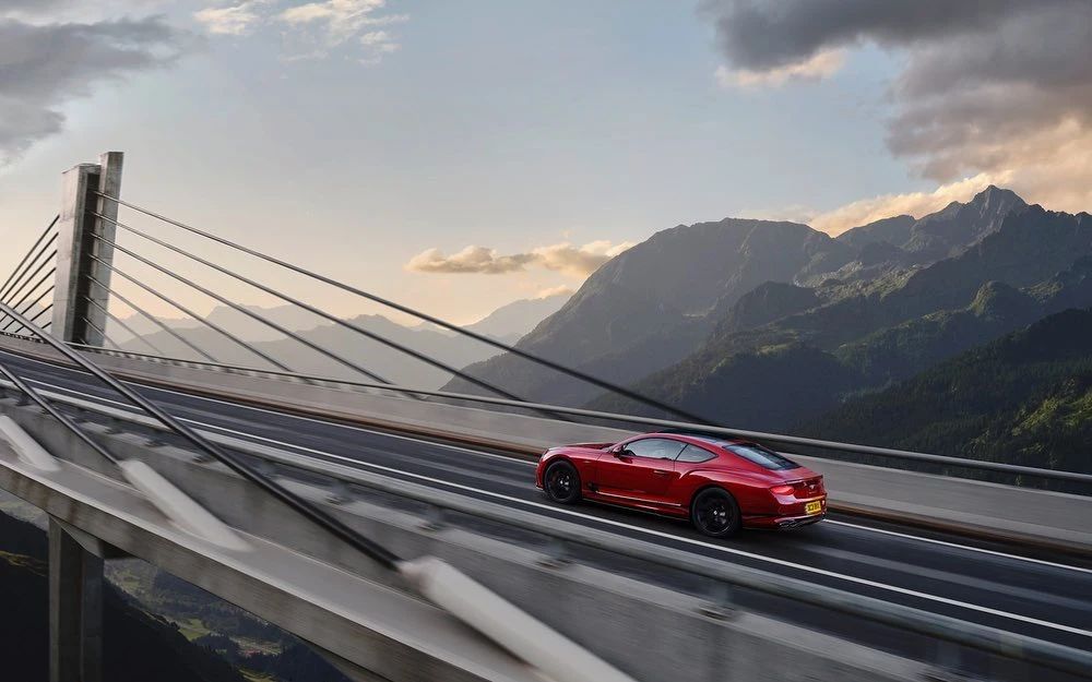 Red Bentley coupe driving across modern suspension bridge with mountain range backdrop under a clear blue, late afternoon sky.