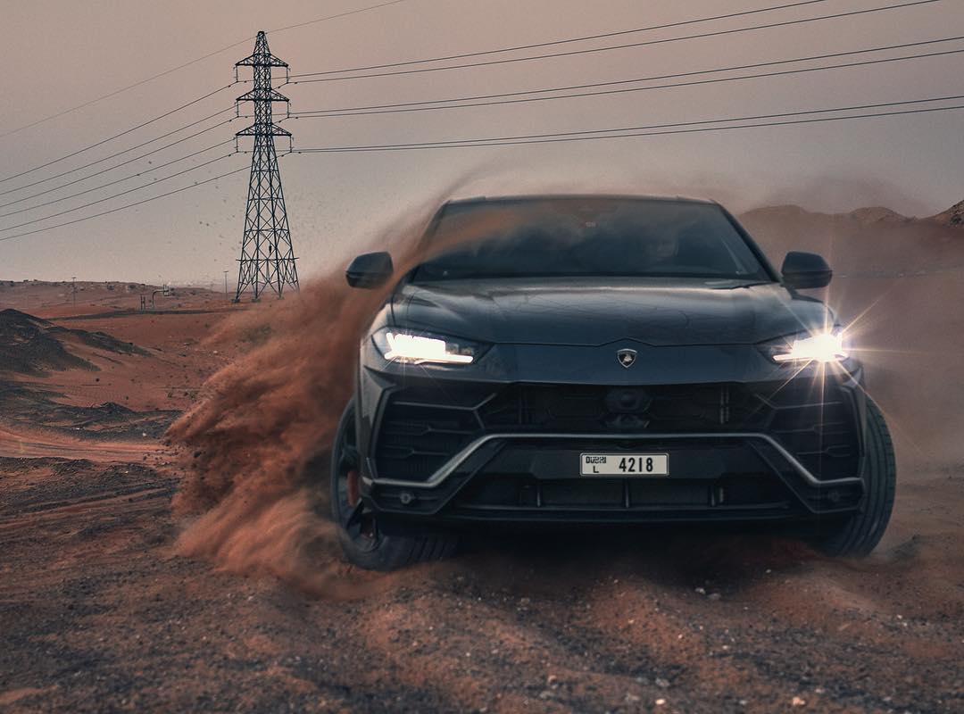Black Lamborghini Urus SUV driving through a desert landscape with sand blowing, power lines in background, automotive photography.