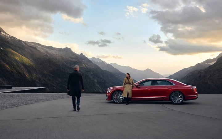 A red Bentley sedan is parked on a grey concrete overlook with two people and a mountain landscape under a cloudy sky in the background.