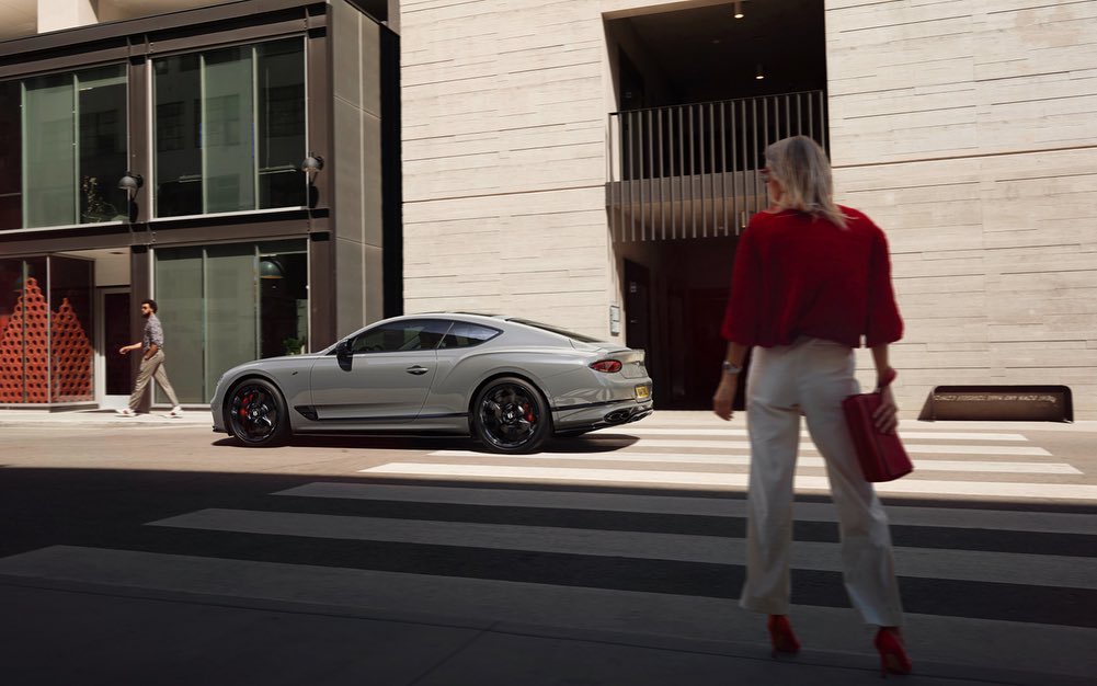 Luxury grey Bentley coupe on an urban street with pedestrians and modern architecture in the background under bright daylight.