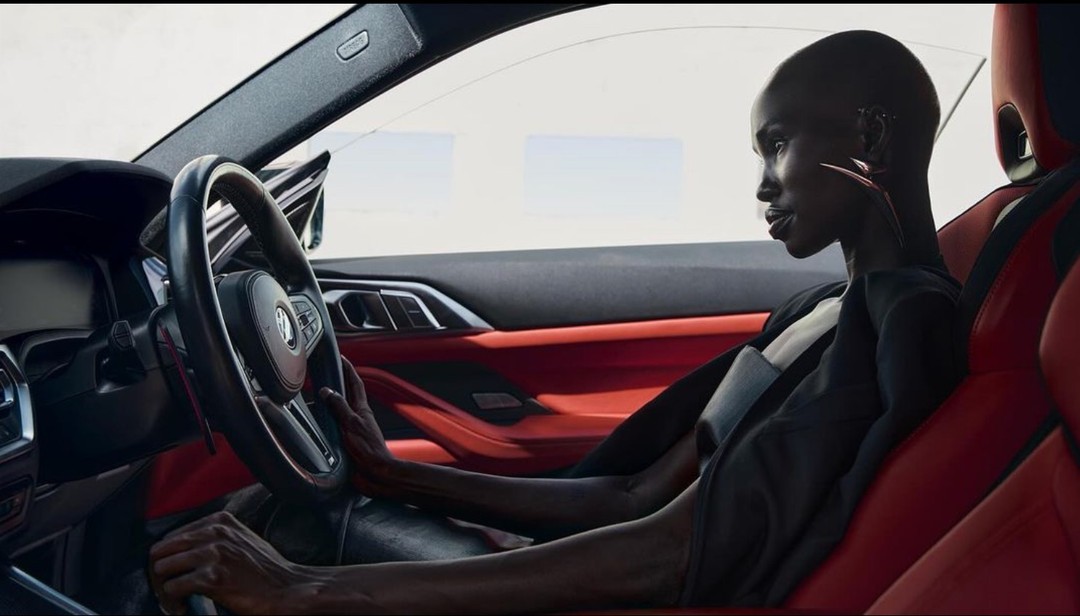 Adult woman sits inside a car with red interior, holding the steering wheel. Automotive fashion shot in natural lighting.